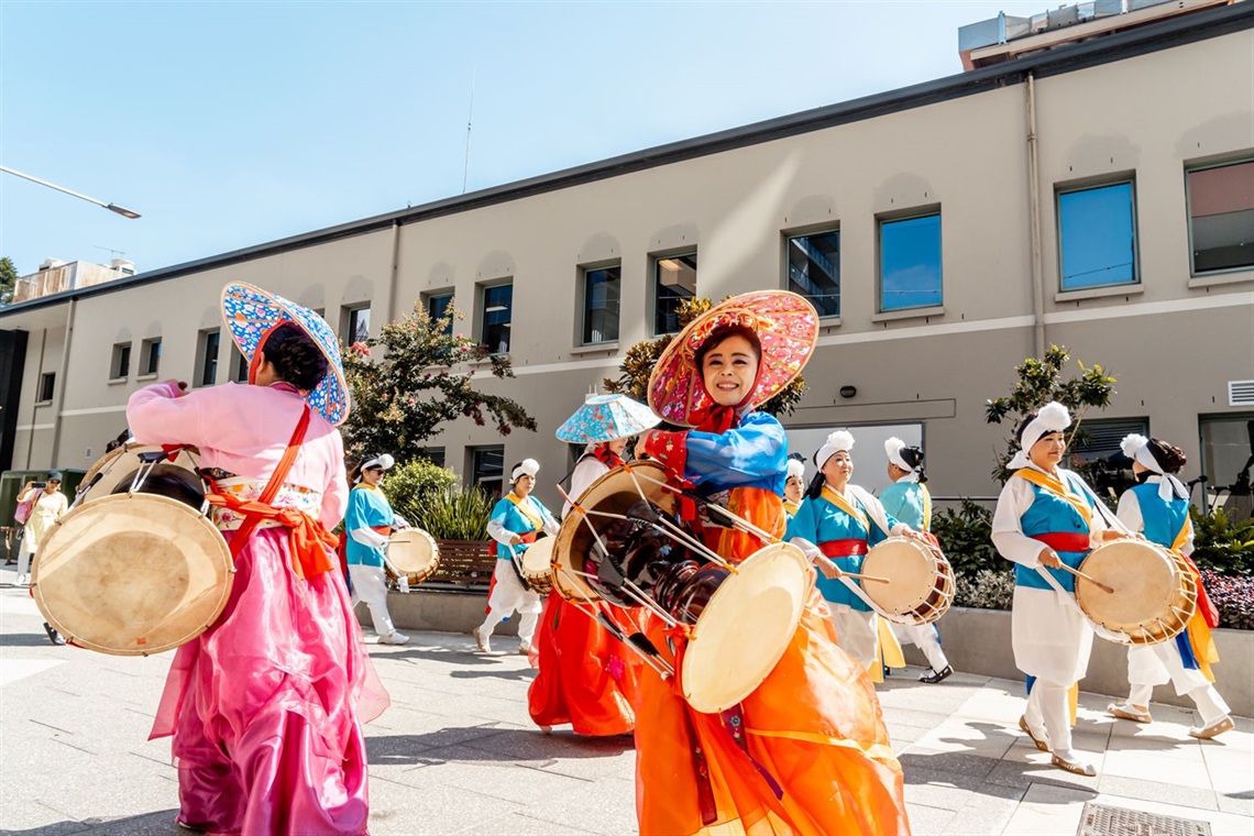 An Asian lady wearing traditional dress playing a large drum in the sun