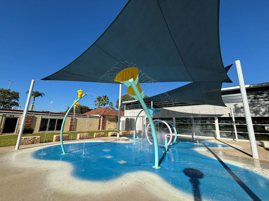 Outdoor splash park on a sunny day. The splash pad is a blue rubberized surface with several colorful water features, including a looping ring and a large yellow bucket that is tipping water. The area is partially shaded by a large gray shade sail, and there are several large stone blocks on the grassy area surrounding the park.