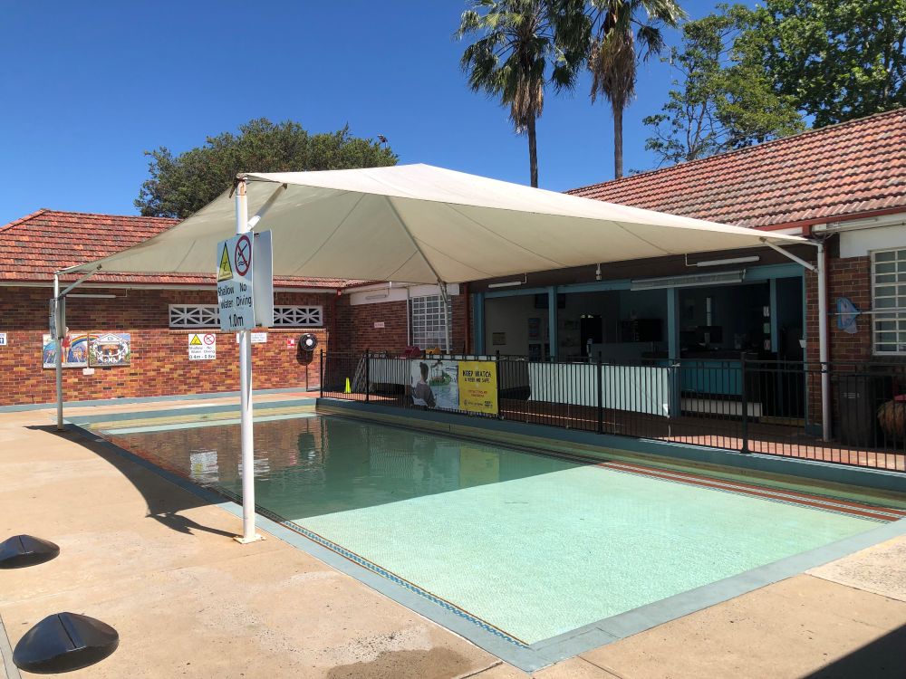 A small, shallow rectangular wading pool with a blue tiled border, situated under a large white shade cloth. The pool is surrounded by a brick building with a reddish tile roof and palm trees are visible in the background against a clear blue sky. A yellow sign next to the pool reads SHALLOW END, NO DIVING, 1.0m